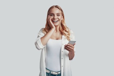 Akıllı telefon kullanan ve gri arka planda dururken gülümseyen çekici genç kadın.