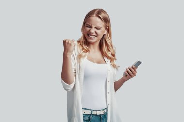 Akıllı telefon kullanan ve gri arka planda dururken gülümseyen çekici genç kadın.