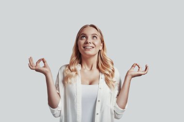 Gri arka planda dururken meditasyon yapan ve gülümseyen güzel genç bir kadın.