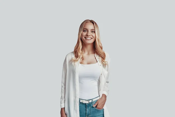 Atractiva joven mujer mirando a la cámara y sonriendo mientras está de pie sobre un fondo gris — Foto de Stock