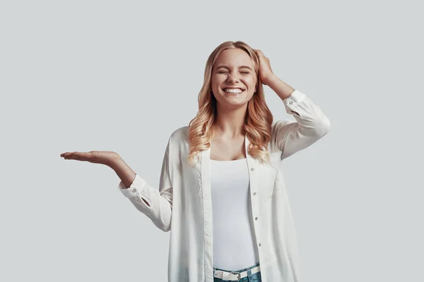 Hermosa mujer joven manteniendo los ojos cerrados y apuntando espacio de copia mientras está de pie sobre el fondo gris — Foto de Stock