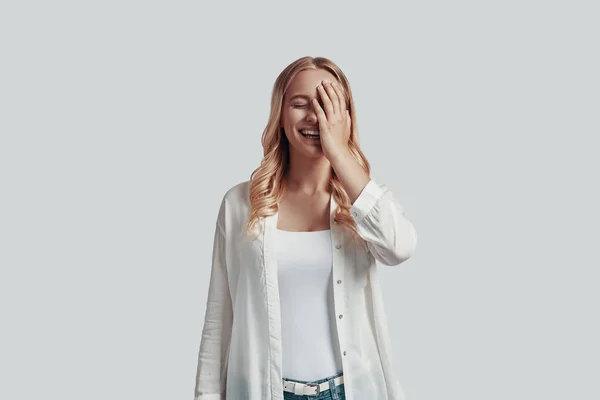 Beautiful young woman covering face with hand and smiling while standing against grey background — Stock Photo, Image