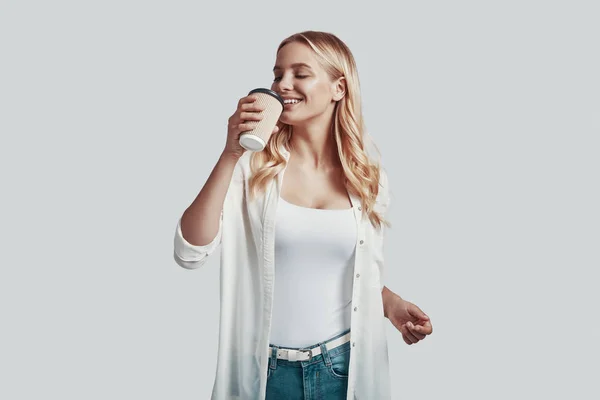 Atractiva joven bebiendo café y sonriendo mientras está de pie sobre fondo gris — Foto de Stock