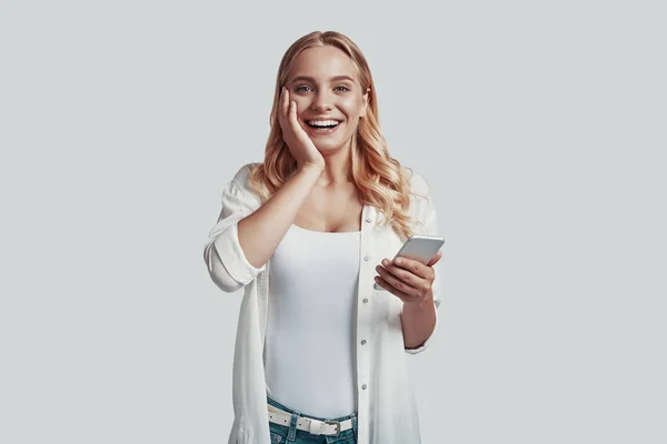 Atrakcyjna młoda kobieta korzystająca ze smartfona i uśmiechnięta stojąc na szarym tle — Zdjęcie stockowe