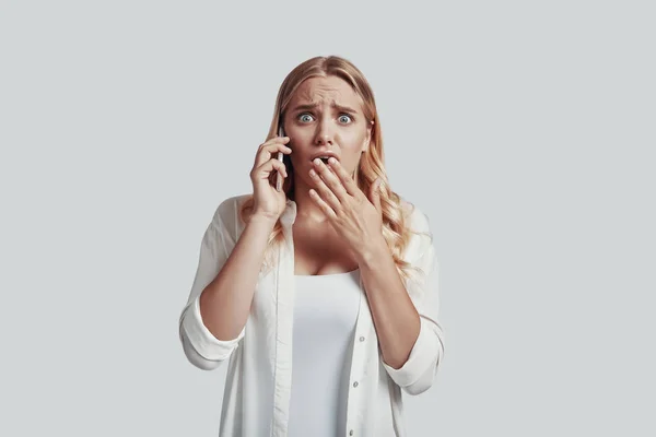 Frustrada jovem mulher falando no telefone inteligente e fazendo um rosto enquanto está de pé contra fundo cinza — Fotografia de Stock
