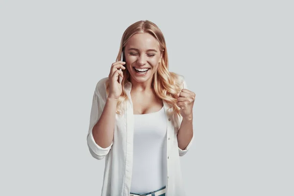 Mujer joven atractiva hablando en el teléfono inteligente y sonriendo mientras está de pie sobre fondo gris —  Fotos de Stock