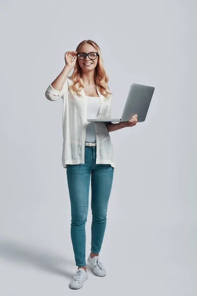 Longitud completa de atractiva mujer joven ajustando las gafas y el uso de la computadora portátil, mientras que de pie contra el fondo gris —  Fotos de Stock