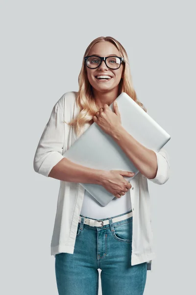 Atractiva joven en gafas que lleva portátil y sonríe mientras está de pie sobre fondo gris —  Fotos de Stock