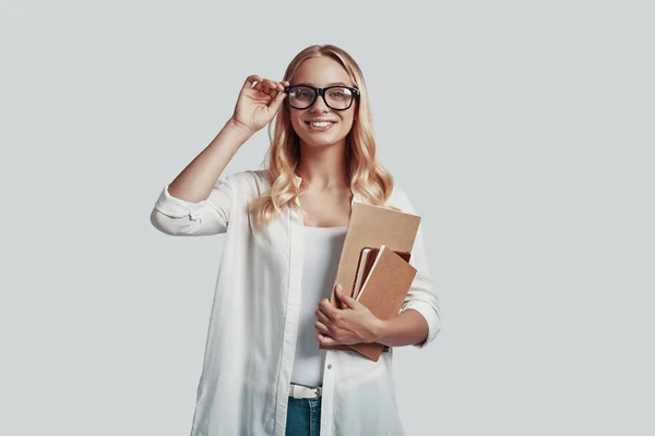 Atractiva joven en gafas que lleva libros y sonríe mientras está de pie sobre fondo gris —  Fotos de Stock