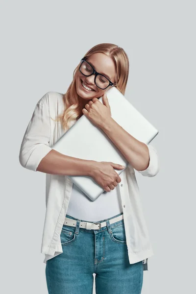 Atractiva joven en gafas que lleva portátil y sonríe mientras está de pie sobre fondo gris —  Fotos de Stock