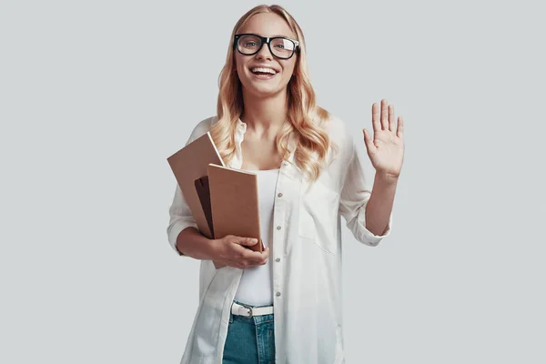 Atractiva joven en gafas que lleva libros y sonríe mientras está de pie sobre fondo gris —  Fotos de Stock