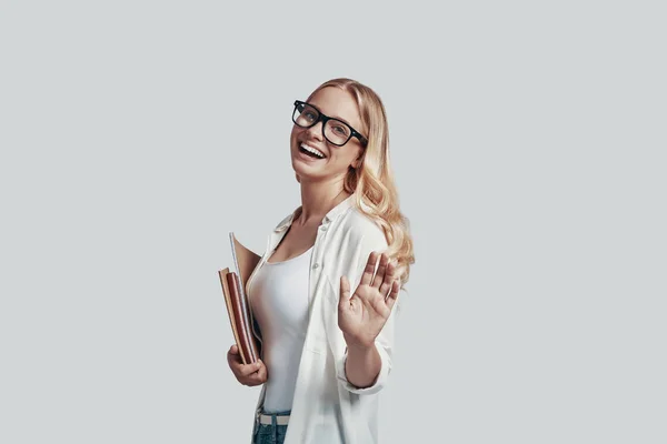 Atractiva joven en gafas que lleva libros y sonríe mientras está de pie sobre fondo gris —  Fotos de Stock