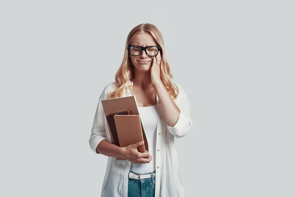 Mujer joven frustrada en gafas llevando libros y haciendo una cara mientras está de pie sobre fondo gris —  Fotos de Stock