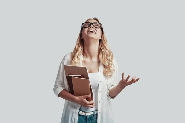 Mujer joven furiosa en gafas llevando libros y gritando mientras está de pie sobre fondo gris —  Fotos de Stock