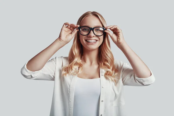 Atractiva joven mujer ajustando gafas y sonriendo mientras está de pie sobre fondo gris —  Fotos de Stock