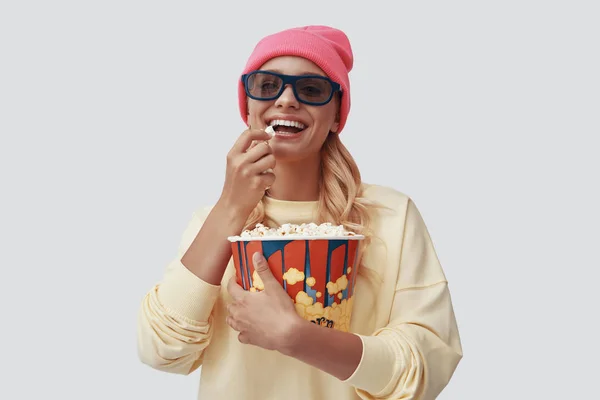 Attraente giovane donna mangiare popcorn e sorridere mentre in piedi contro sfondo grigio — Foto Stock