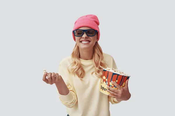 Atractiva joven mujer comiendo palomitas de maíz y sonriendo mientras está de pie sobre fondo gris —  Fotos de Stock