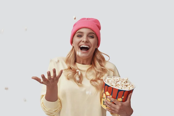 Attraente giovane donna gettando popcorn e sorridente mentre in piedi contro sfondo grigio — Foto Stock
