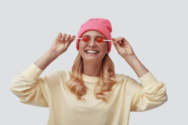 Mujer joven atractiva sonriendo y cubriendo los ojos con piruletas mientras está de pie sobre fondo gris —  Fotos de Stock