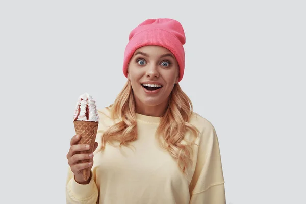 Atractiva joven mujer comiendo helado y mirando a la cámara mientras está de pie sobre un fondo gris —  Fotos de Stock