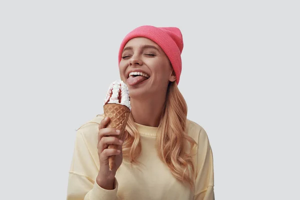 Ελκυστική νεαρή γυναίκα τρώει παγωτό και κάνει γκριμάτσα ενώ στέκεται στο γκρίζο φόντο — Φωτογραφία Αρχείου