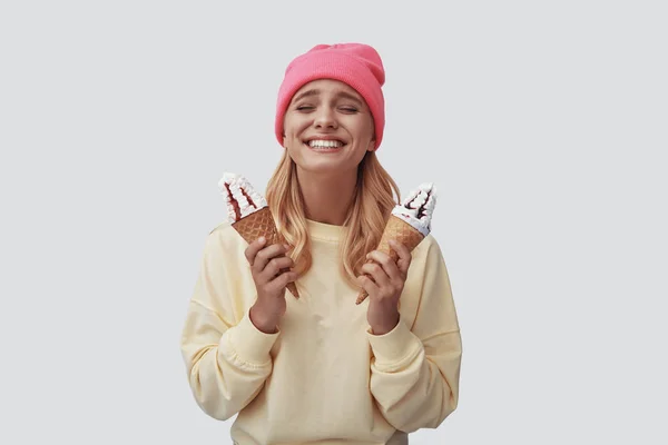 Attractive young woman eating ice cream and making a face while standing against grey background — Stock Photo, Image