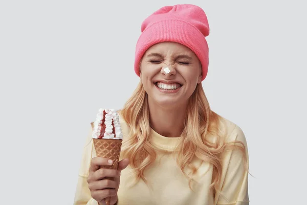Ελκυστική νεαρή γυναίκα τρώει παγωτό και χαμογελά ενώ στέκεται πάνω σε γκρι φόντο — Φωτογραφία Αρχείου