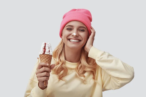Attraente giovane donna mangiare gelato e guardando la fotocamera mentre in piedi sullo sfondo grigio — Foto Stock