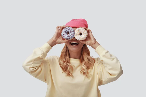 Jovem atraente cobrindo os olhos com donuts e sorrindo enquanto está de pé contra fundo cinza — Fotografia de Stock