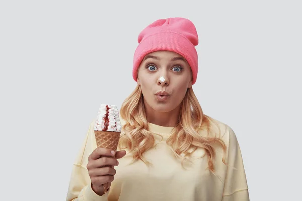 Attractive young woman eating ice cream and looking at camera while standing against grey background — Stock Photo, Image