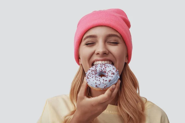 Jovem atraente comendo donut e sorrindo de pé contra fundo cinza — Fotografia de Stock