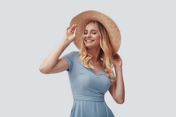 Mujer joven atractiva sonriendo y ajustando su sombrero mientras está de pie sobre fondo gris —  Fotos de Stock