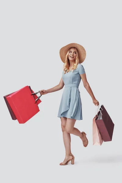 Full length of attractive young woman smiling and carrying shopping bags while standing against grey background — Stock Photo, Image