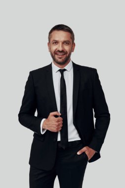 Yakışıklı, takım elbiseli, gülümseyen ve gri arka planda kameraya bakan genç bir adam.