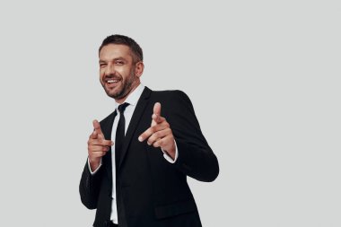 Yakışıklı, takım elbiseli, gülümseyen ve gri arka planda dururken sana işaret eden genç bir adam.