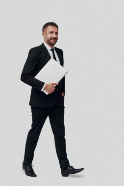 Tam takım elbiseli yakışıklı genç bir adam dizüstü bilgisayar taşıyor ve gri arka planda yürürken gülümsüyor.