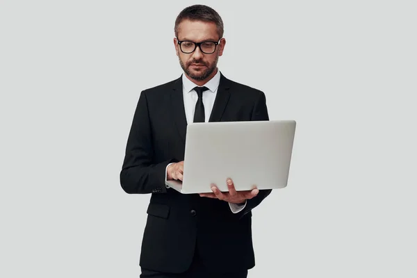 Jovem encantador em terno completo usando laptop enquanto está de pé contra fundo cinza — Fotografia de Stock