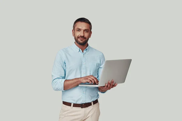 Jovem bonito usando laptop e sorrindo enquanto está de pé contra fundo cinza — Fotografia de Stock