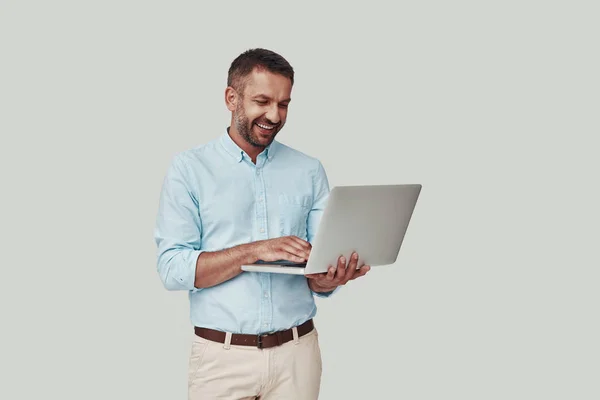 Jovem bonito usando laptop e sorrindo enquanto está de pé contra fundo cinza — Fotografia de Stock