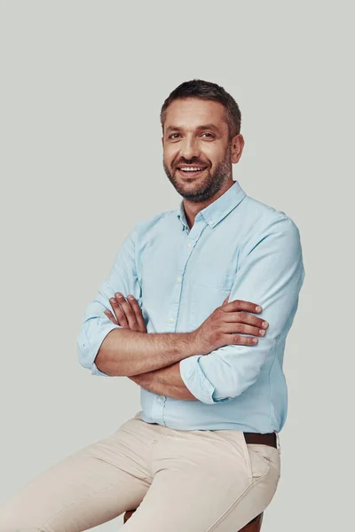 Beau jeune homme regardant la caméra et souriant tout en étant assis sur fond gris — Photo