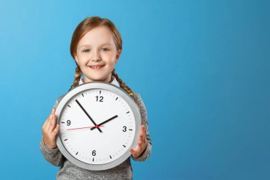 Mavi bir arka plan üzerinde büyük bir saat tutan sevimli bir küçük kız. Eğitim kavramı, okul, zamanlama, öğrenme zamanı. Kopyalama alanı.