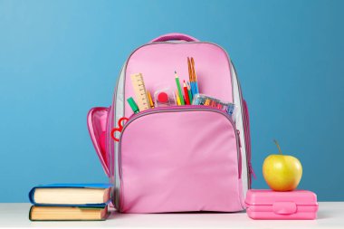 Öğrenci seti. Kırtasiyeli pembe sırt çantası, bir yığın kitap, bir beslenme çantası, mavi arka plan üzerinde masanın üzerinde bir elma. Okula geri dön.