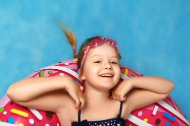 Mayo giymiş küçük bir kız, çörek şişme çemberinin üzerinde yatıyordu. Bir çocuğun yakın çekim portresi. Mavi arka plan. Üstte görüntü.