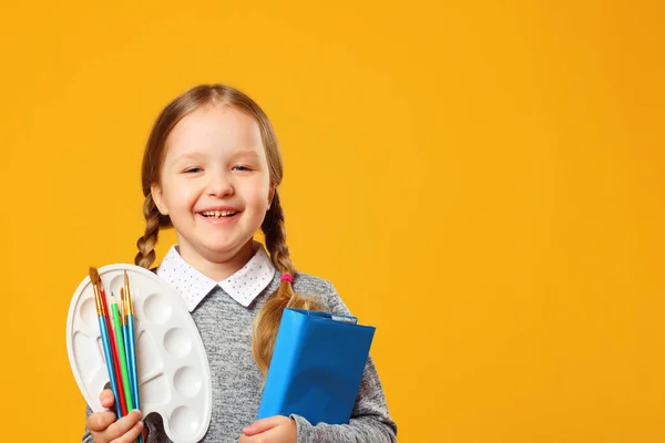 Sarı bir arka plan üzerinde neşeli bir küçük kız portresi. Schoolgirl bir kitap, kalem, fırça ve bir palet tutar. Eğitim kavramı. Kopyalama alanı. — Stok fotoğraf