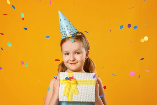 Cheerful little girl celebrates birthday. A child holds a box with a gift in the rain of confetti. Closeup portrait on yellow background. — Stock Photo, Image