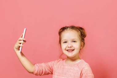Pembe bir arka plan üzerinde saç çörekler ile sevimli bir küçük kız Closeup portre. Çocuk telefonu tutar ve selfie çeker
