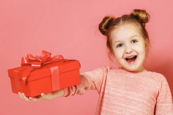 Ritratto ravvicinato di una graziosa bambina con panini sui capelli su sfondo rosa. Il bambino tiene fuori una scatola con un regalo . — Foto Stock