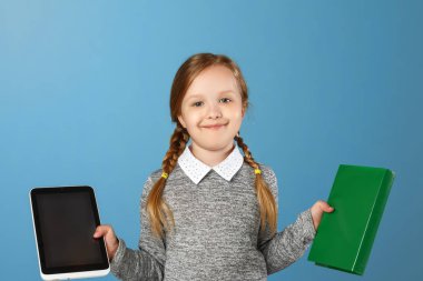 Mavi bir arka planda neşeli küçük kız. Çocuk bir kitap ve tablet tutar. Eğitim kavramı, okul, seçim, karşılaştırma.