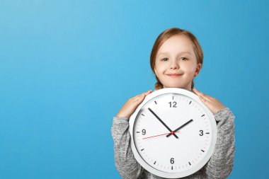 Mavi bir arka plan üzerinde büyük bir saat tutan sevimli bir küçük kız. Eğitim kavramı, okul, zamanlama, öğrenme zamanı. Kopyalama alanı.