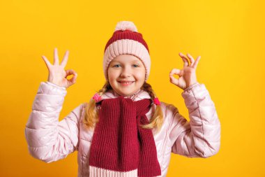 Sarı arka planda ceketli, atkılı ve şapkalı küçük bir kız. Çocuk iki eliyle ok işareti gösterir. Sonbahar, başarı ve eğitim kavramı.